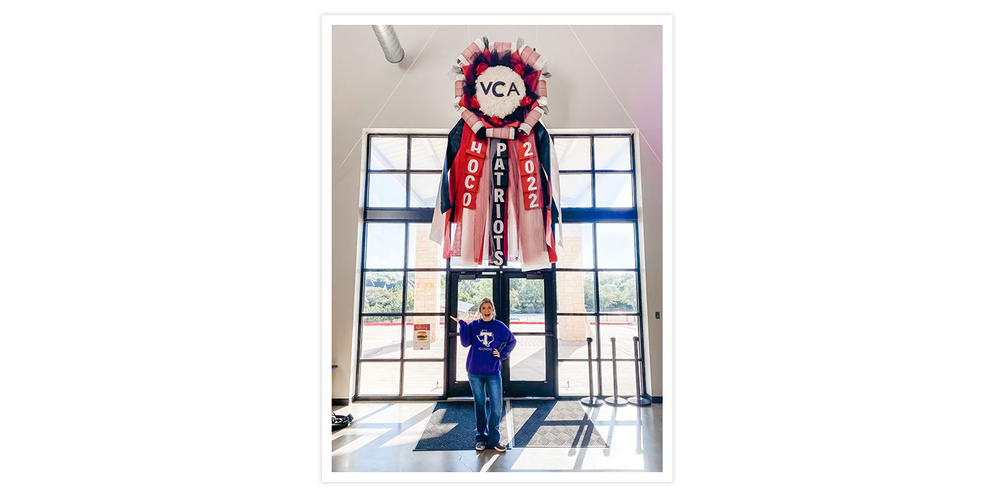 A woman with blonde hair holding her hand in the air wearing a purple Tarleton State University sweatshirt, posing with a giant Homecoming mum hanging on a wall.